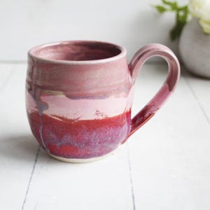 May include: A handmade ceramic mug with a rounded body and a large handle. The mug features a gradient of colors, transitioning from a deep red at the base to lighter pinks and purples towards the rim. The mug is designed for hot beverages.