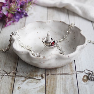 May include: A speckled, off-white ceramic ring dish with a raised center for rings. A silver ring with a red gemstone sits on the ring holder. A silver necklace with floral accents and a silver chain are draped in the dish.
