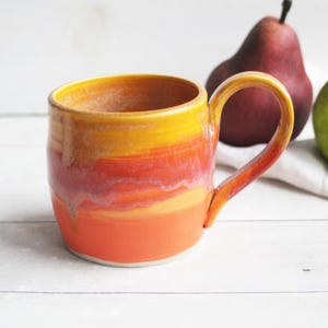 May include: A ceramic mug featuring a vibrant gradient glaze that transitions from yellow at the rim to orange and red tones toward the base. The mug has a rounded body and a curved handle on the right side, showcasing a smooth, glossy finish.