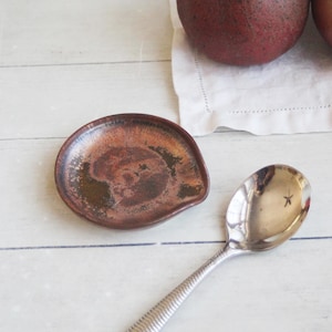 May include: A small, round, dark brown ceramic spoon rest with a mottled, metallic-toned glaze featuring copper and black hues, positioned next to a polished silver-toned metal spoon with a textured, ribbed handle design.