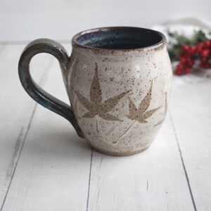 May include: A handmade ceramic mug with a speckled, off-white finish. The mug features brown maple leaf designs and a dark blue rim and handle. The mug is a unique piece of pottery, perfect for enjoying coffee or tea.