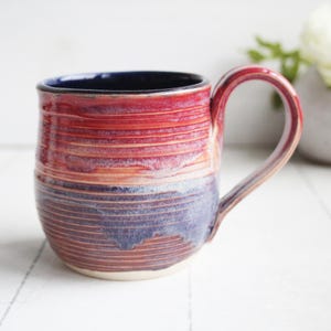 May include: A handmade ceramic mug with a red, orange, and blue color scheme. The mug features a rounded body with a handle and a dark blue interior. The exterior has a textured, horizontal striped pattern.