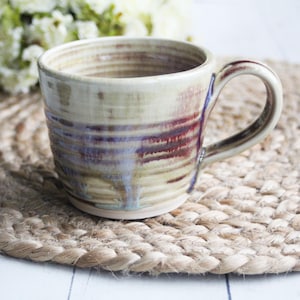 May include: A handmade ceramic mug with a light brown glaze and a red and purple stripe design. The mug has a rounded shape and a handle.