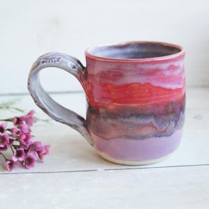 May include: A ceramic mug with a glossy glaze featuring horizontal bands of pink, red, and purple. The mug has a curved handle with a textured, greyish-purple finish and a smooth, rounded rim. The base is a light, neutral cream color.