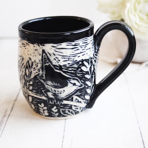 May include: Black and white ceramic mug featuring a detailed illustration of a bird perched on a branch amidst foliage. The mug has a glossy black interior and handle, with a contrasting white exterior design. A unique and artistic piece for enjoying beverages.