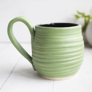 May include: A handmade green ceramic mug with a textured, ridged design. The mug has a curved handle and a black interior. The base of the mug is a light cream color. The mug is a light green color.