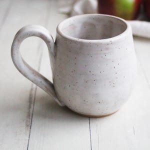 May include: A speckled white ceramic mug with a rounded body and a large, curved handle. The mug has a slightly textured surface with small brown speckles. The mug is sitting on a white wooden surface.