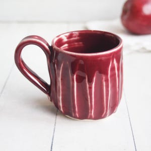 May include: A handmade, burgundy-colored ceramic mug with a textured, ridged exterior. The mug has a curved handle and a white base. The glossy glaze gives the mug a handcrafted look. A red apple is visible in the background.