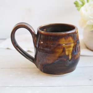 May include: A ceramic mug with a rounded body and a curved handle, featuring a glossy glaze in shades of deep brown, amber, and hints of dark blue. The mug has a smooth, reflective surface with visible color variations and a white base.