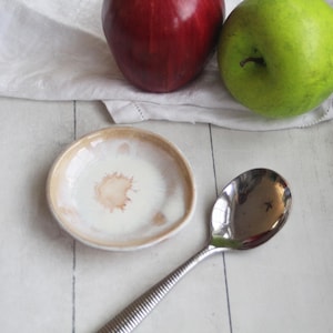 May include: A white ceramic spoon rest with a brown splatter design. The spoon rest is sitting on a white wooden surface. A silver spoon is lying on the surface next to the spoon rest. A red apple and a green apple are in the background.