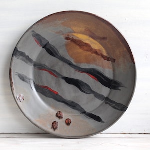 May include: A large, round ceramic plate with a gray glaze and abstract black, red, and brown painted designs. The plate has a slightly raised rim.