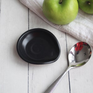 May include: A small, black ceramic spoon rest with a smooth, glossy finish. The spoon rest is oval-shaped and sits on a white wooden surface, next to a stainless steel spoon and two green apples.