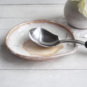 May include: A ceramic spoon rest with a round, shallow shape, featuring a cream-colored glaze with a prominent tan-brown center and a matching tan-brown rim. A stainless steel cooking spoon with a black handle rests inside the ceramic dish.
