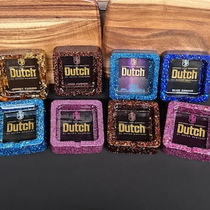 May include: Eight square ashtrays with glitter finishes. Each ashtray features a gold logo with the text "Dutch" and a flavor name. The flavors are Honey Fusion, Java Fusion, Blue Dream, and an unreadable flavor.