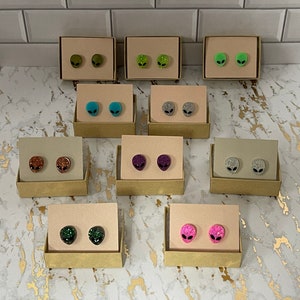May include: Assortment of alien head stud earrings displayed on small cardboard boxes. The earrings are made of resin and have glitter embedded in them. The colors of the earrings include green, blue, silver, brown, purple, black, and pink.