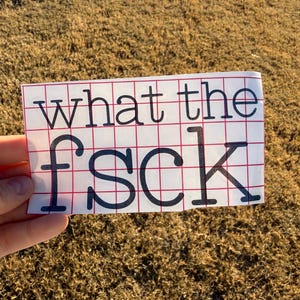 May include: A white sticker with a red grid pattern and the words "what the f*ck" in black, held against a brown background.