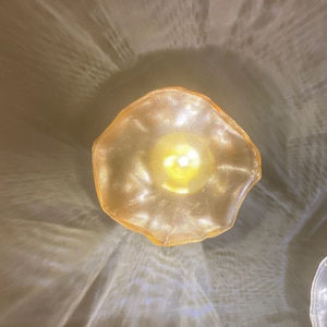 May include: A circular, golden-hued pendant light fixture with a textured, mesh-like surface. The light emits a warm, glowing illumination from the center, casting intricate, rippling patterns of light and shadow onto the surrounding area.