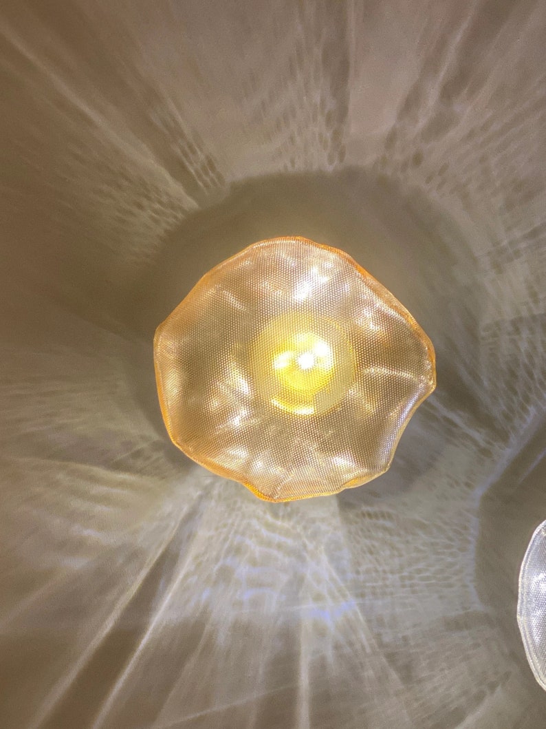 May include: A circular, golden-hued pendant light fixture with a textured, mesh-like surface. The light emits a warm, glowing illumination from the center, casting intricate, rippling patterns of light and shadow onto the surrounding area.