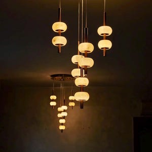 May include: A multi-pendant ceiling light fixture featuring two circular black mounts. Each mount supports several thin vertical rods with glowing, translucent, off-white disc-shaped shades arranged in a tiered, cascading formation.