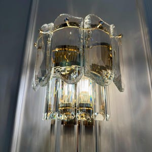 May include: A vintage crystal wall sconce with a gold-toned metal frame. The sconce features multiple clear crystal elements, including rectangular and faceted shapes, and is designed to hold multiple light bulbs. The sconce is mounted on a light gray wall.