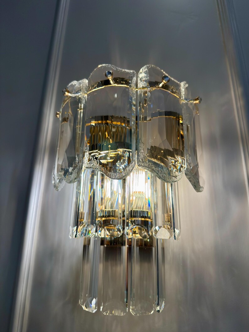 May include: A vintage crystal wall sconce with a gold-toned metal frame. The sconce features multiple clear crystal elements, including rectangular and faceted shapes, and is designed to hold multiple light bulbs. The sconce is mounted on a light gray wall.