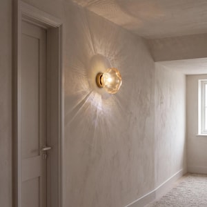 May include: A wall-mounted light fixture featuring a round, amber-tinted glass shade with a textured, organic surface. The lamp is attached to a circular brass base and emits a warm, glowing light that creates a sunburst pattern on the wall.