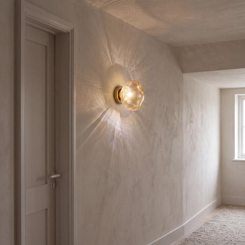 May include: A wall-mounted light fixture featuring a round, amber-tinted glass shade with a textured, organic surface. The lamp is attached to a circular brass base and emits a warm, glowing light that creates a sunburst pattern on the wall.