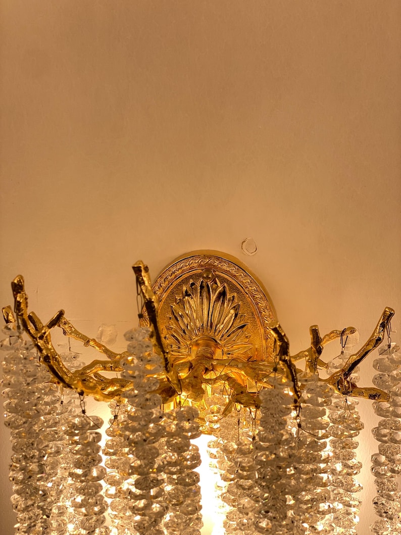 May include: A gold-toned wall sconce with a crystal chandelier design. The sconce features a decorative, ornate, circular backplate with a floral design. The sconce has a branch-like design with cascading crystal beads.