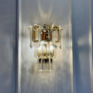 May include: A crystal wall sconce with a gold-toned metal frame. The sconce features multiple tiers of clear, rectangular crystals and a central amber-colored crystal element. The light fixture is mounted on a white wall.