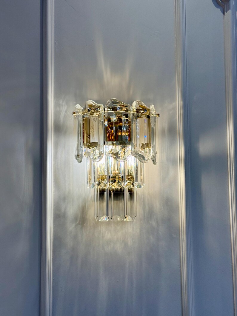 May include: A crystal wall sconce with a gold-toned metal frame. The sconce features multiple tiers of clear, rectangular crystals and a central amber-colored crystal element. The light fixture is mounted on a white wall.