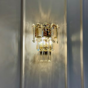 May include: A wall-mounted light fixture with a gold-toned metal frame and clear crystal accents. The fixture has multiple tiers of rectangular and teardrop-shaped crystals, creating a sparkling effect. The light casts a pattern on the wall.