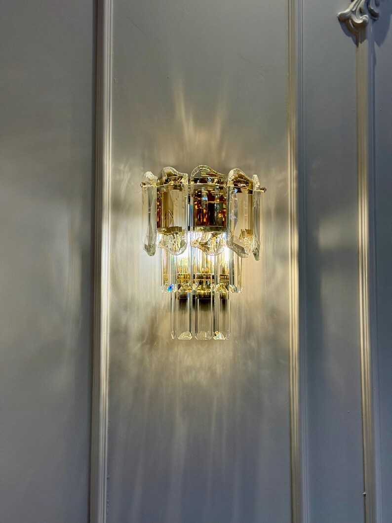 May include: A wall-mounted light fixture with a gold-toned metal frame and clear crystal accents. The fixture has multiple tiers of rectangular and teardrop-shaped crystals, creating a sparkling effect. The light casts a pattern on the wall.