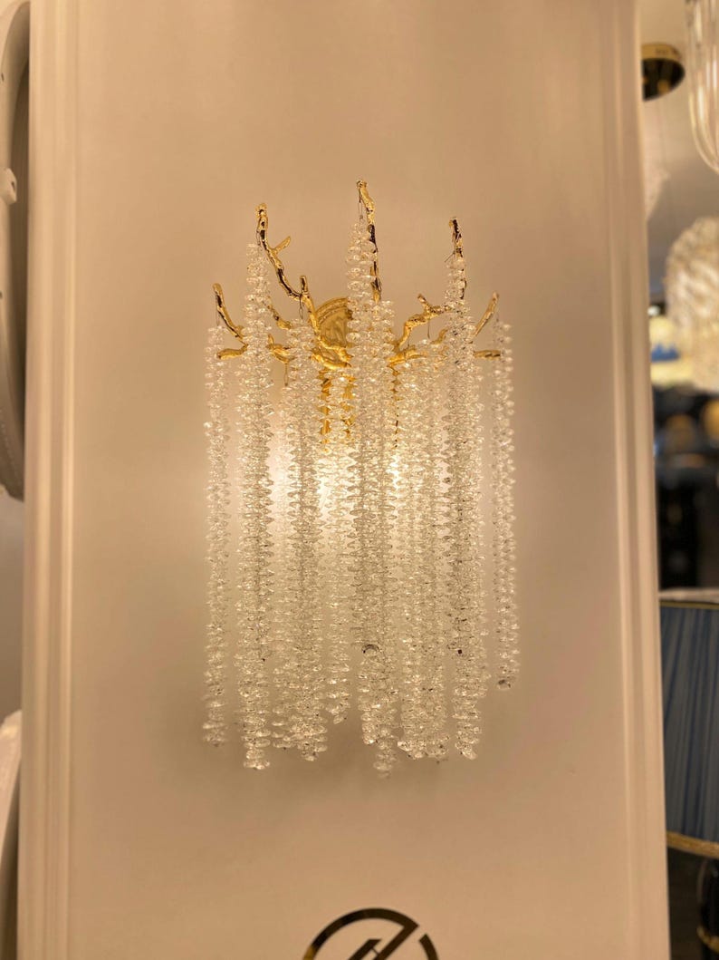 May include: A gold wall sconce with a crystal chandelier design. The sconce features a branch-like frame with cascading crystal beads.