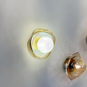 K&ouml;nnte beinhalten: Zwei Wandleuchten mit bernsteinfarbenen, organisch geformten Glasschirmen. Eine Leuchte enth&auml;lt eine hellwei&szlig;e, kugelf&ouml;rmige Gl&uuml;hbirne, w&auml;hrend die andere eine warm leuchtende Filamentbirne enth&auml;lt, die beide Licht abgeben.