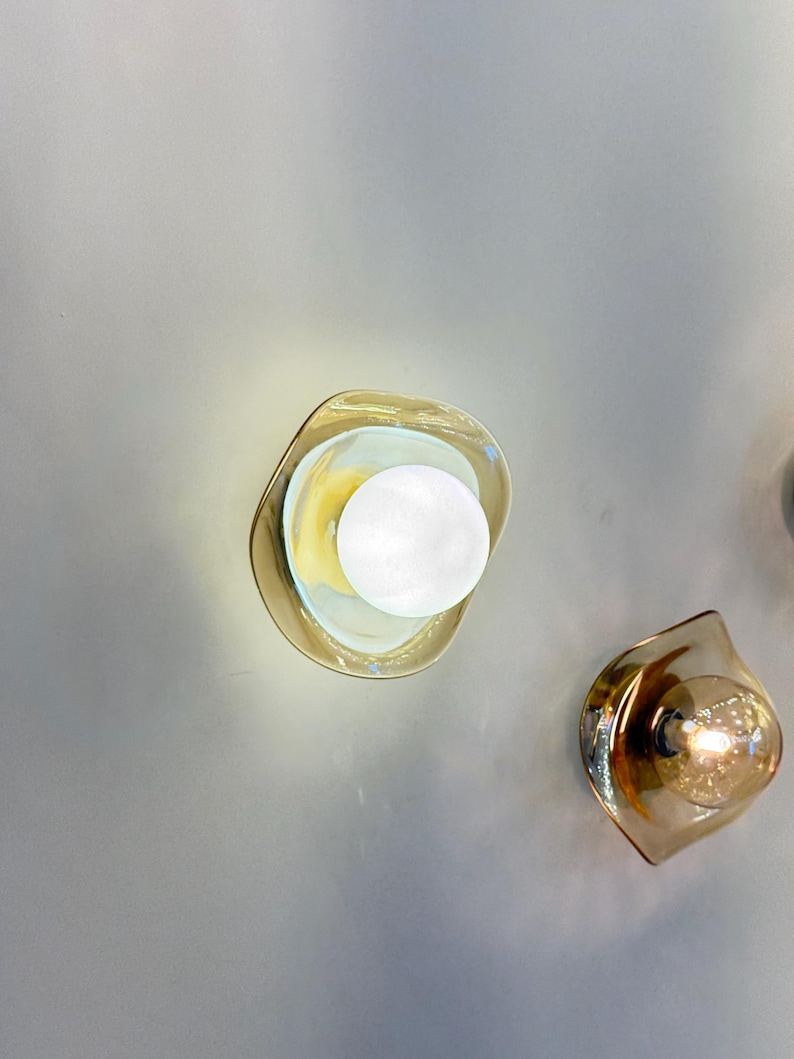 K&ouml;nnte beinhalten: Zwei Wandleuchten mit bernsteinfarbenen, organisch geformten Glasschirmen. Eine Leuchte enth&auml;lt eine hellwei&szlig;e, kugelf&ouml;rmige Gl&uuml;hbirne, w&auml;hrend die andere eine warm leuchtende Filamentbirne enth&auml;lt, die beide Licht abgeben.