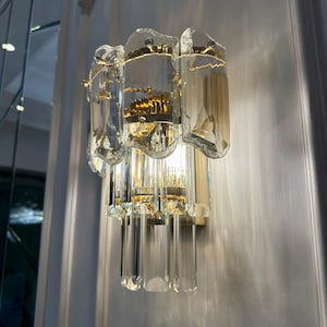 May include: A wall-mounted light fixture with a gold-toned metal base and clear, faceted crystal accents. The fixture has a layered design with rectangular and elongated crystal elements, creating a sparkling effect. The light is on, casting shadows on the wall.