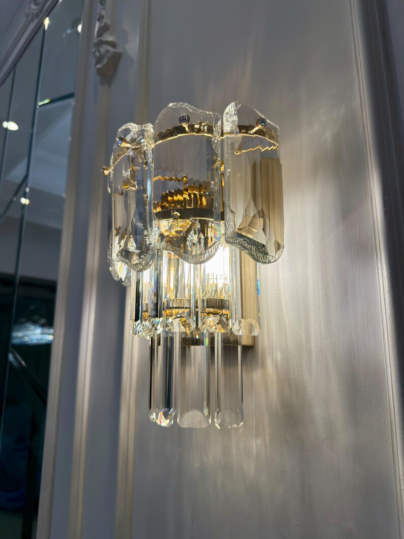 May include: A wall-mounted light fixture with a gold-toned metal base and clear, faceted crystal accents. The fixture has a layered design with rectangular and elongated crystal elements, creating a sparkling effect. The light is on, casting shadows on the wall.