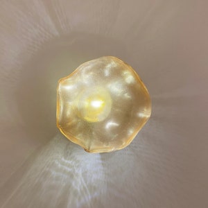 May include: A circular, gold-toned metal mesh lampshade with an irregular, wavy edge. The shade features a fine, perforated grid pattern that allows a bright, warm light to shine through from the center, casting soft, diffused light patterns.