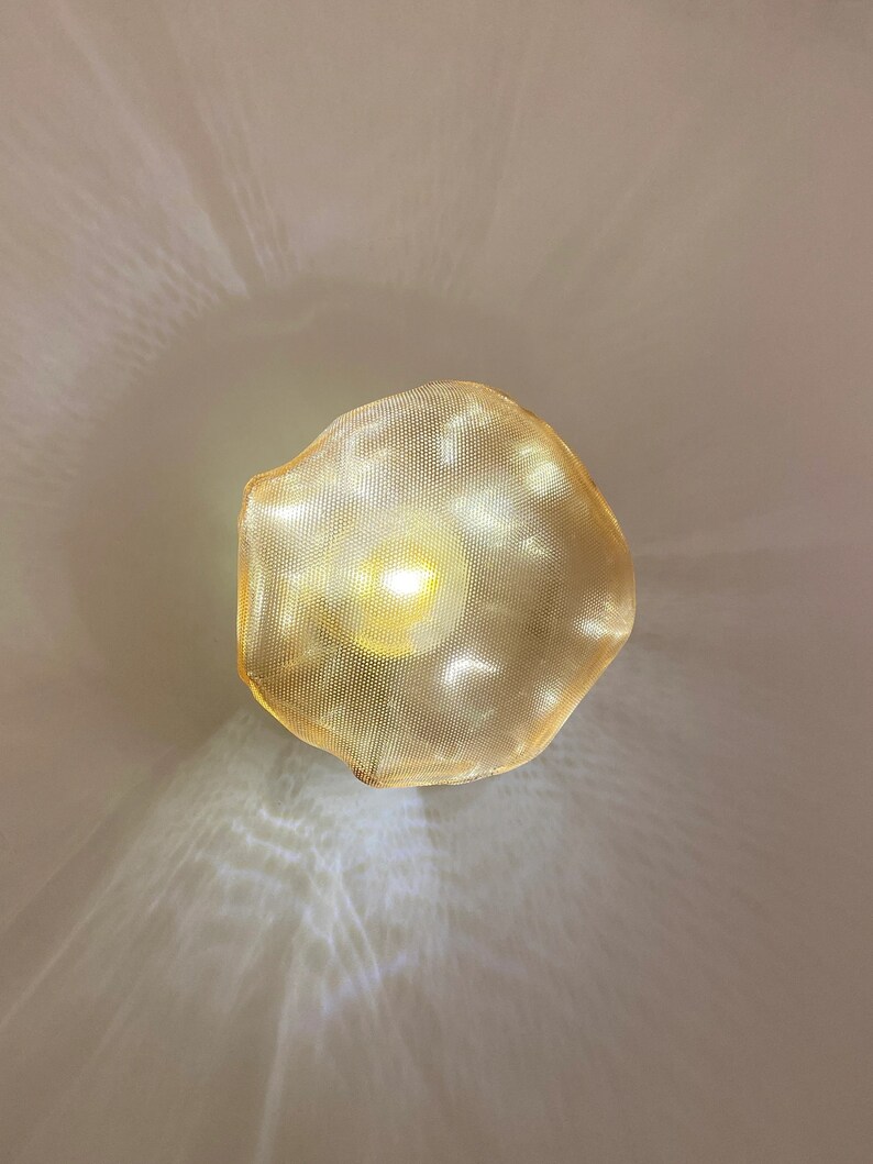 May include: A circular, gold-toned metal mesh lampshade with an irregular, wavy edge. The shade features a fine, perforated grid pattern that allows a bright, warm light to shine through from the center, casting soft, diffused light patterns.
