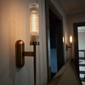 May include: A wall-mounted light fixture featuring a cylindrical, vertically ribbed glass shade held by a brass-toned metal base. The fixture has a long, slender, tapered metal stem extending downward from the light source.