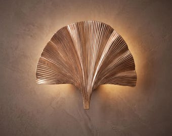 May include: A wall-mounted light fitting shaped like a ginkgo leaf with a metallic bronze finish. The surface features fine, radiating vertical ridges that catch the light, creating a warm glow that emanates from behind the fan-shaped structure.