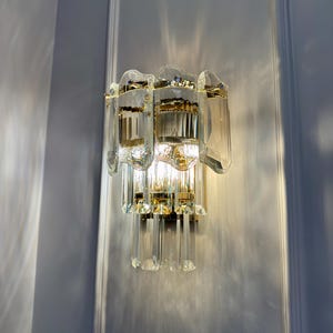 May include: A wall-mounted light fixture with a gold-colored metal frame and clear, faceted glass crystals. The fixture has a layered design with multiple tiers of glass elements, creating a sparkling effect when illuminated. The light is turned on, casting a warm glow.