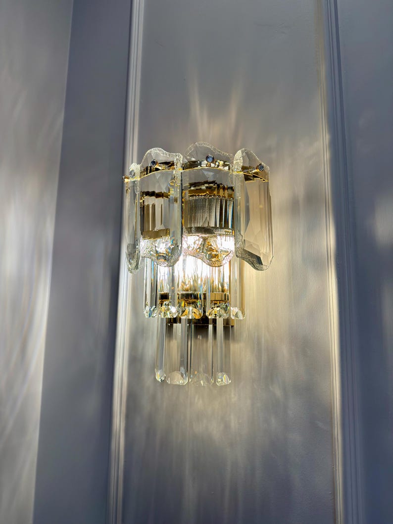 May include: A wall-mounted light fixture with a gold-colored metal frame and clear, faceted glass crystals. The fixture has a layered design with multiple tiers of glass elements, creating a sparkling effect when illuminated. The light is turned on, casting a warm glow.