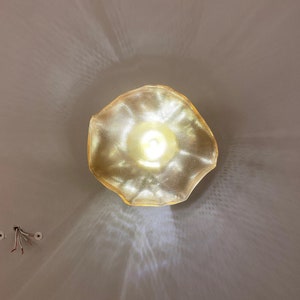 May include: A gold-toned, textured pendant light fixture with an irregular, organic, wavy-edged shade. The light emits a bright, warm glow, casting intricate, radiating patterns of light and shadow onto the surrounding surface.