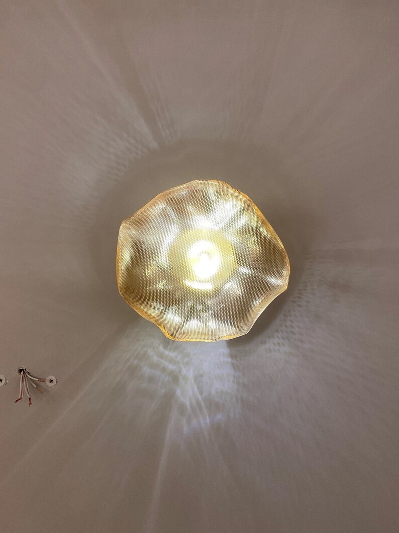 May include: A gold-toned, textured pendant light fixture with an irregular, organic, wavy-edged shade. The light emits a bright, warm glow, casting intricate, radiating patterns of light and shadow onto the surrounding surface.