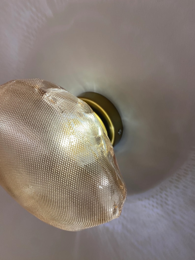 May include: A wall-mounted light fixture featuring a textured, amber-tinted glass shade with a fine dot pattern. The shade is attached to a circular, matte brass-colored metal base that secures the light to the wall surface.