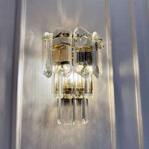 May include: A wall-mounted light fixture with a gold-colored metal frame and clear crystal accents. The fixture has multiple tiers of crystal rods and a decorative top. The light is on, casting shadows on the white wall.