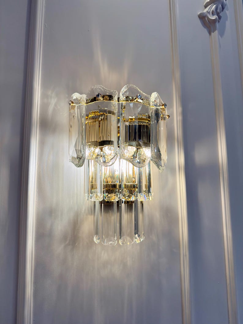 May include: A wall-mounted light fixture with a gold-colored metal frame and clear crystal accents. The fixture has multiple tiers of crystal rods and a decorative top. The light is on, casting shadows on the white wall.