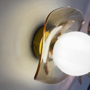 K&ouml;nnte beinhalten: Eine Wandleuchte mit einem runden Messingsockel und einem durchscheinenden, bernsteinfarbenen Glasschirm mit gewelltem, organischem Rand. Eine kugelf&ouml;rmige, undurchsichtige wei&szlig;e Gl&uuml;hbirne ist mittig im Schirm platziert und strahlt hell.