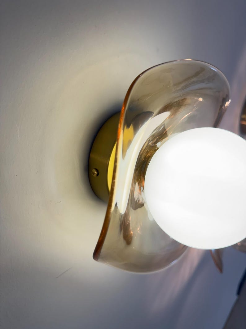 K&ouml;nnte beinhalten: Eine Wandleuchte mit einem runden Messingsockel und einem durchscheinenden, bernsteinfarbenen Glasschirm mit gewelltem, organischem Rand. Eine kugelf&ouml;rmige, undurchsichtige wei&szlig;e Gl&uuml;hbirne ist mittig im Schirm platziert und strahlt hell.